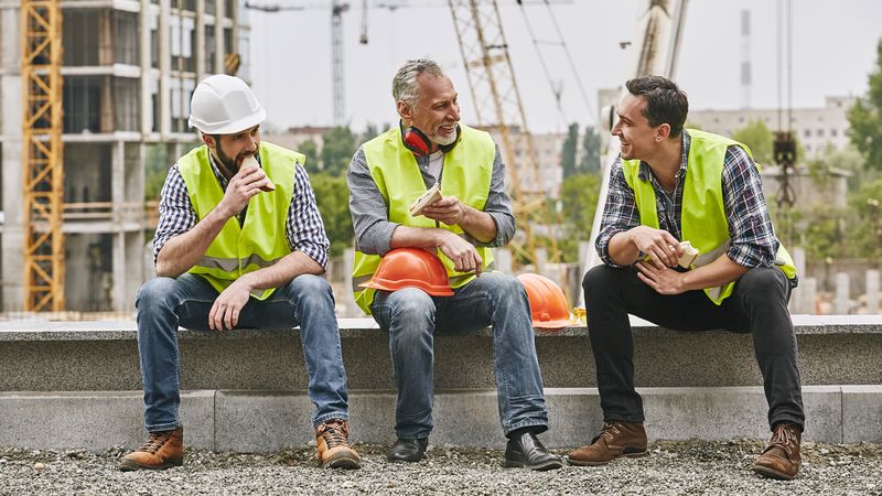 Three construction workers sitting down on site eating sandwiches