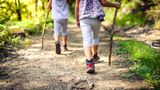 Two young children walking along a woodland footpath surrounded by trees. They are wearing hiking boots and holding large sticks 