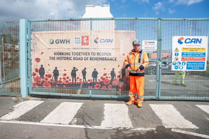 Peter Osborne in high-visibility clothing stands beside a construction site gate with a banner reading &ldquo;Working together to reopen the historic Road of Remembrance,&rdquo; alongside safety signage.