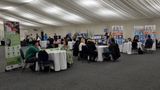A large room with different companies set up at stalls scattered around the room. Parents sit at tables talking to professionals 