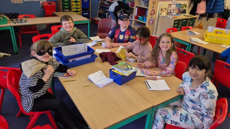 Six children sitting around a table in their classroom in fancy dress reading books and taking part in craft activities 