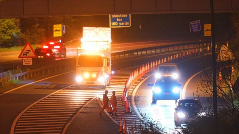 Night-time roadworks on a dual carriageway, with traffic cones narrowing lanes as workers in high‑visibility clothing cross the road and vehicles pass under bright work lights.