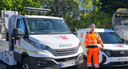 &ldquo;A highway worker in high‑visibility orange clothing and safety helmet stands beside two white Kent County Council vehicles on a tree‑lined roadside, with maintenance equipment visible on the trucks.&rdquo;