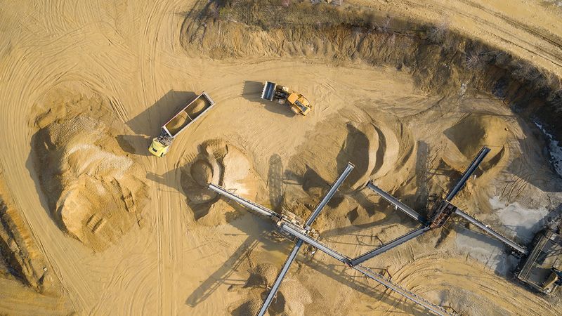 Aerial view of a sand and gravel quarry showing conveyor belts, large stockpiles of aggregate, a dump truck transporting material, and a loader operating on sandy terrain. Vehicle tracks and excavation areas are visible across the site.