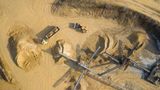 Aerial view of a sand and gravel quarry showing conveyor belts, large stockpiles of aggregate, a dump truck transporting material, and a loader operating on sandy terrain. Vehicle tracks and excavation areas are visible across the site.