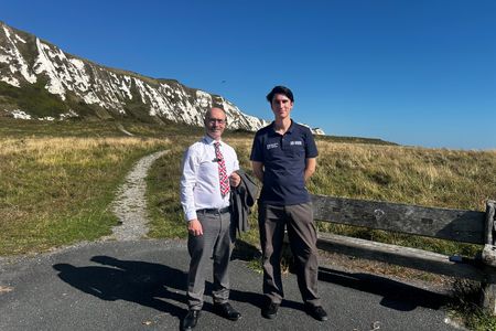 Two people standing beside a coastal path at the foot of white chalk cliffs, with grassland and clear blue sky in the background.