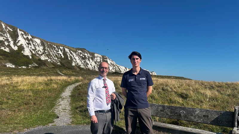 Two people standing beside a coastal path at the foot of white chalk cliffs, with grassland and clear blue sky in the background.