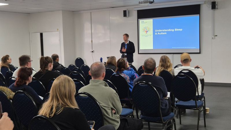 A man standing in front of a projector screen talking to a room full of people sat down in chairs listening