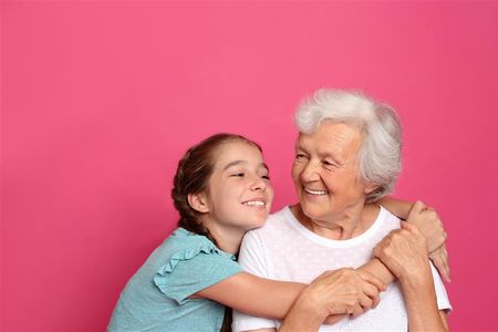  Young girl hugs an older woman as they smile at each other against a pink background