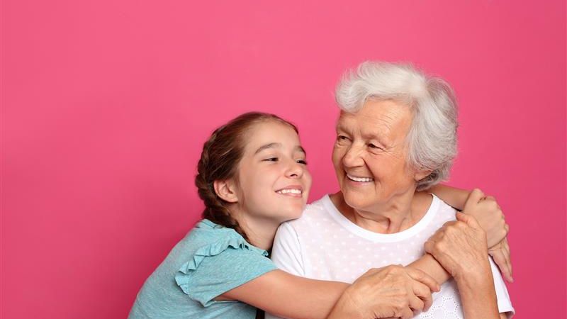  Young girl hugs an older woman as they smile at each other against a pink background