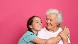  Young girl hugs an older woman as they smile at each other against a pink background