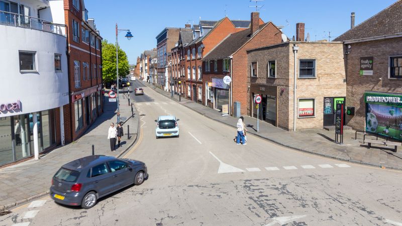 View of the junction connecting St Dunstan&rsquo;s Street and Station Road West in Canterbury, showing a two‑lane road with cars and pedestrians. There building on both sides of the road, a zebra crossing in the background, traffic signs, and a bus stop.
