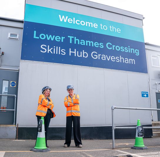 Large exterior sign reading &ldquo;Welcome to the Lower Thames Crossing Skills Hub Gravesham&rdquo; on a grey building, with two people in high-visibility orange jackets and hard hats standing in front near green traffic cones.