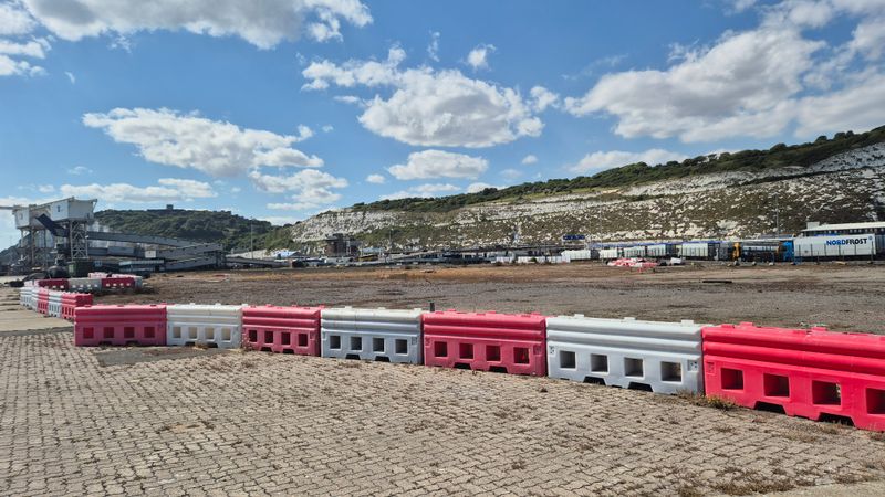 Open area at the Port of Dover with red and white safety barriers in the foreground, cargo handling infrastructure to the left, and the white cliffs of Dover stretching across the background under a blue sky with scattered clouds.