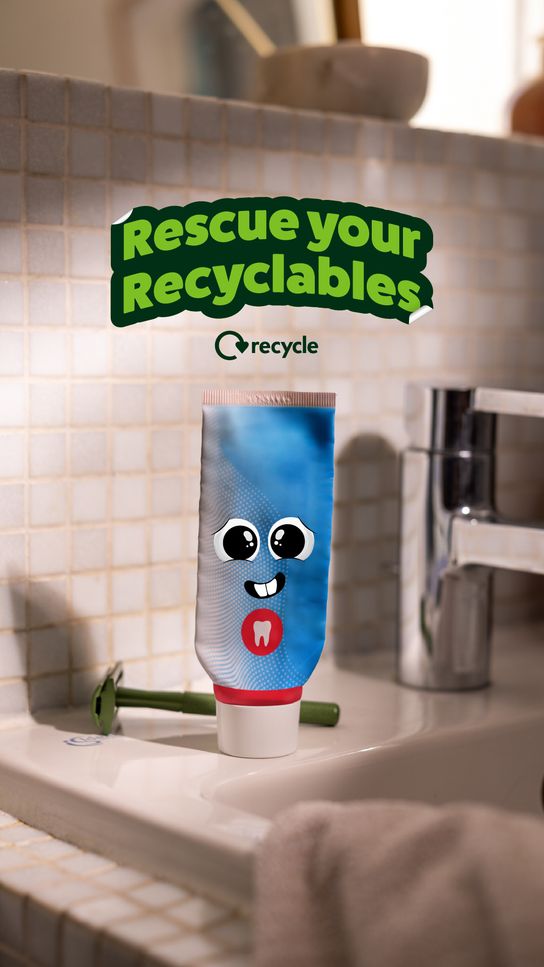 A blue toothpaste tube with a cartoon face and tooth symbol stands upright on a bathroom sink beside a green razor. Above the tube, the text &ldquo;Rescue your recyclables&rdquo; and the Recycle logo appear, promoting the recycling of bathroom items. The sink is set against a tiled wall with a chrome tap in soft, warm lighting.