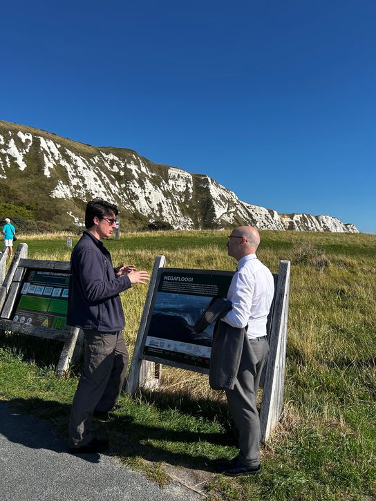 Two people looking at an interpretation board in front of white chalk cliffs and open grassland on the coast.