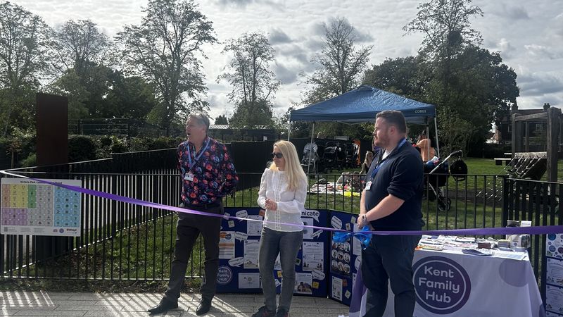 Three people stand in front of a purple ribbon in a park.