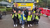 A group of primary school children wearing high-visibility vests standing in front of a Kent County Council gritter lorry named "Grit Britain", with accompanying adults and roadwork barriers visible in the background.
