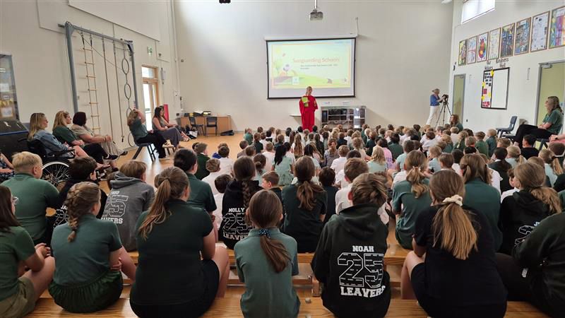 Pupils at Platt Primary School sit on the floor in the school hall for an assembly with Michelle Baker from the Melanoma Fund. A projector shows informative slides on a screen 
