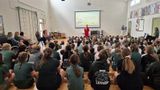 Pupils at Platt Primary School sit on the floor in the school hall for an assembly with Michelle Baker from the Melanoma Fund. A projector shows informative slides on a screen 