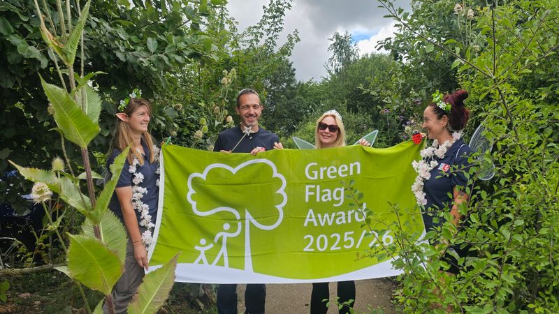 Four people wearing fairy wings stand in the countryside holding a big green flag which says Green Flag awards 25/26.