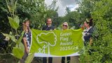Four people wearing fairy wings stand in the countryside holding a big green flag which says Green Flag awards 25/26.