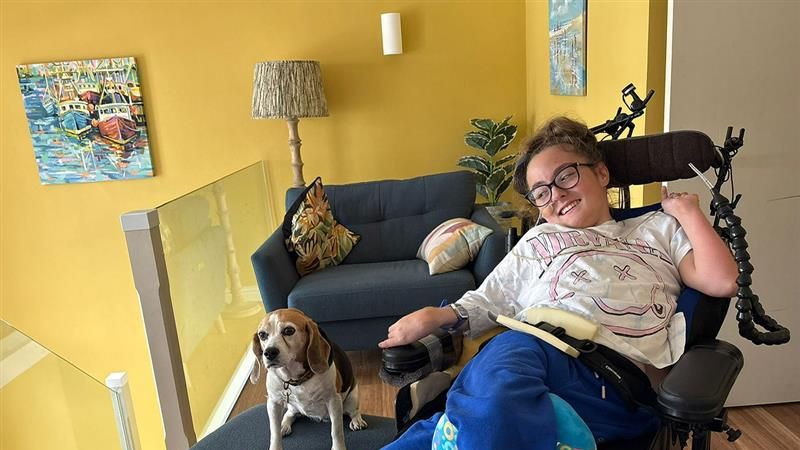 Lucie Lewis in her wheelchair at home next to her beagle Poppy who is sat on a blue sofa stool