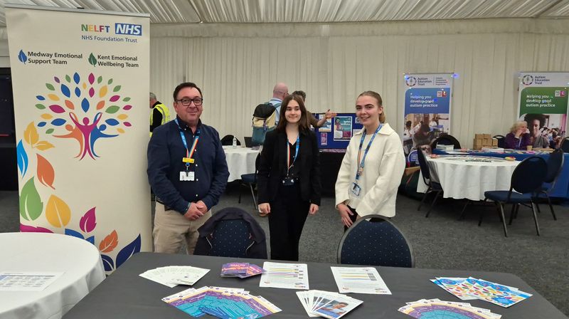 Three members of staff from NELFT wearing landyards stand next to their banner and behind a table full of information about their service