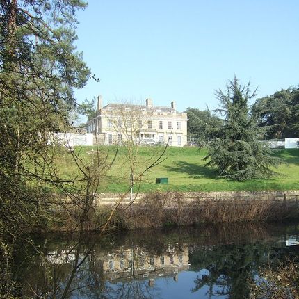 Large historic manor house on a grassy hill with trees, reflected in a still pond in the foreground.