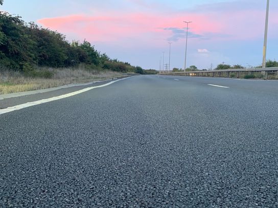 Low-angle view of a newly resurfaced road with visible tarmac texture, an empty carriageway stretching into the distance under a pink and blue evening sky.