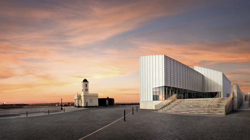 Exterior of Turner Contemporary in Margate at sunset, showing the modern glass‑fronted gallery building, wide steps and the historic clock tower on the seafront.