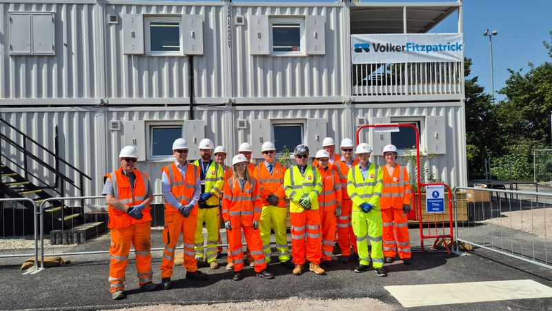 Group of people wearing high-visibility orange and yellow clothing and white hard hats standing in front of stacked portable site cabins, with a &ldquo;VolkerFitzpatrick&rdquo; banner displayed above.