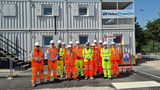 Group of people wearing high-visibility orange and yellow clothing and white hard hats standing in front of stacked portable site cabins, with a &ldquo;VolkerFitzpatrick&rdquo; banner displayed above.