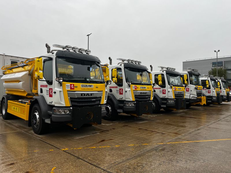 A row of Kent County Council gritter lorries parked at a depot on a wet day, lined up side by side with yellow and white winter maintenance equipment ready for service.