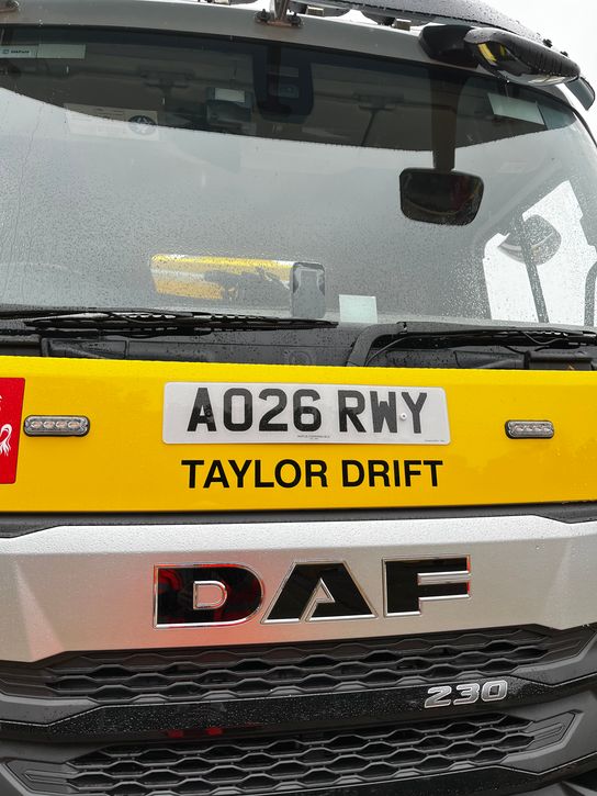 Front view of a yellow Kent gritter lorry on a wet day, with raindrops visible on the windscreen. The vehicle displays the registration plate "AO26 RWY" and the name "TAYLOR DRIFT" across the front above the large DAF logo. The close-up image highlights the gritter's cab and front grille.