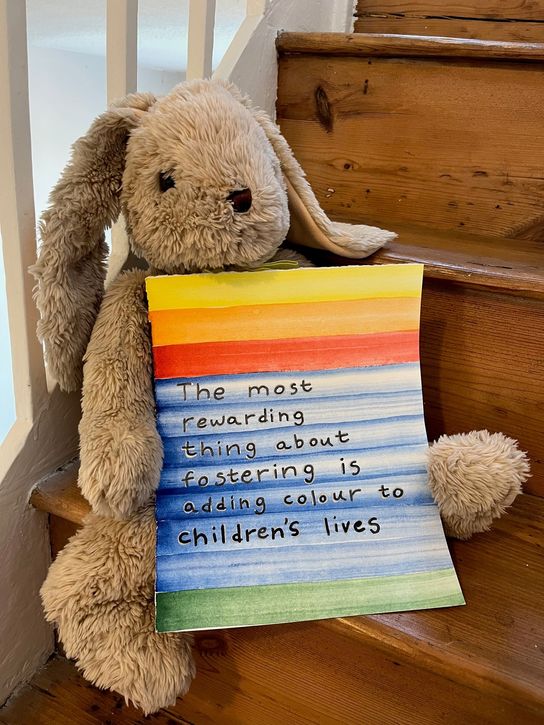 A toy bunny sits on a wooden stair holding a rainbow covered note that says 'The most rewarding thing about fostering is adding colour to children's lives'
