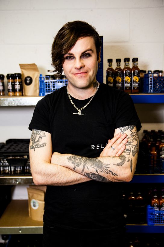 Kier Kemp, who started the Condimaniacs brand in Kent, is seen in front of his stock on shelving. He stands with his arms folded and is wearing a black branded T-shirt