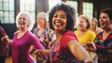 A group of older women, smiling and wearing brightly-coloured t-shirts, enjoy a zumba class