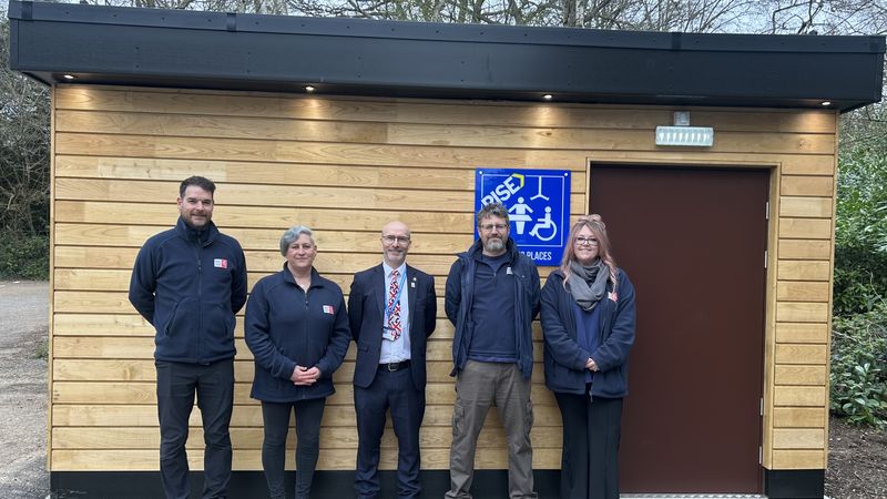 Five people standing in front of a wooden accessible toilet building at Trosley Country Park, with a blue &lsquo;Changing Places&rsquo; sign displayed on the wall.