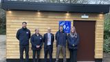 Five people standing in front of a wooden accessible toilet building at Trosley Country Park, with a blue &lsquo;Changing Places&rsquo; sign displayed on the wall.