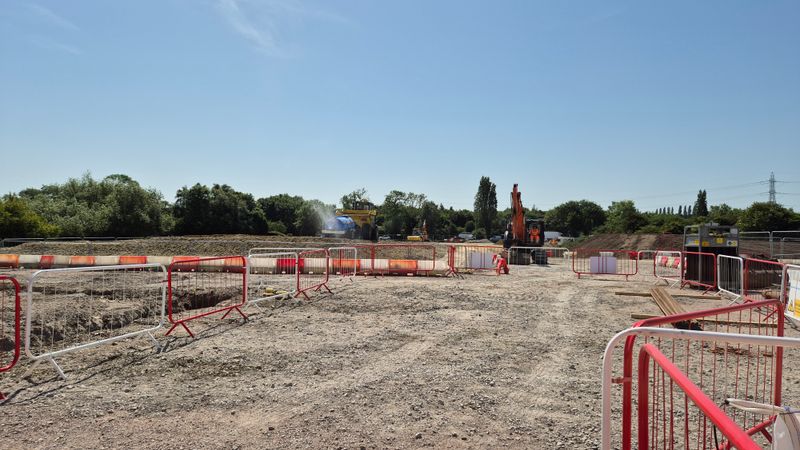 Construction site with gravel ground and red-and-white safety barriers in the foreground, with an excavator and water tanker working on a raised earth embankment in the background under a clear blue sky.