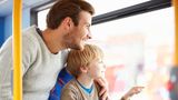 A seated adult and a young child ride on a bus, looking out through a large window. The child points toward something outside while the adult has an arm around the child. Bright natural light fills the bus interior, with blue patterned seats and yellow handrails visible.