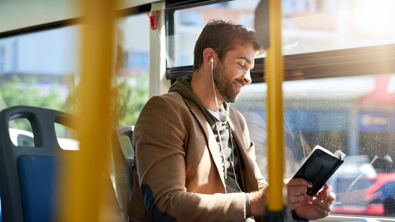 Person seated on a bus, wearing earphones and a casual jacket, reading a notebook or journal by a sunlit window. Yellow handrails frame the foreground, with bus seats and a blurred streetscape visible in the background.