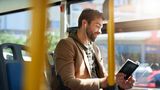 Person seated on a bus, wearing earphones and a casual jacket, reading a notebook or journal by a sunlit window. Yellow handrails frame the foreground, with bus seats and a blurred streetscape visible in the background.