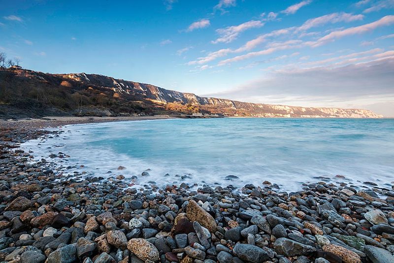 Pebble beach and calm sea beneath chalk cliffs at the Warren, with blue sky and scattered clouds overhead.
