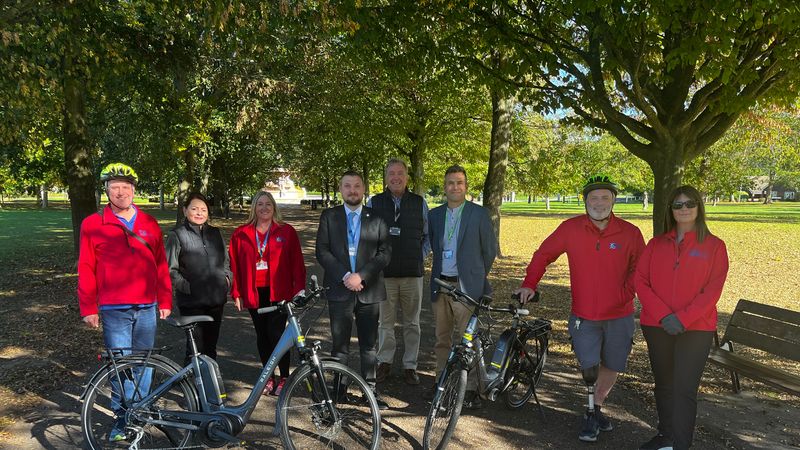 A group of eight people stand on a tree-lined park pathway beside two electric bicycles. Several people are wearing red jackets and cycling helmets. Sunlight filters through the trees, casting shadows across the path, with benches and open green space visible in the background.