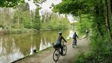 Three people on bikes ride through greenery next to a river.