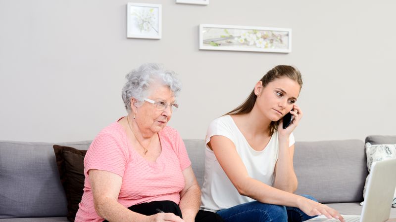 A younger woman and an older woman sit on a sofa with a laptop open on a coffee table in front of them. The younger woman is making a phone call. It looks like they are dealing with some paperwork