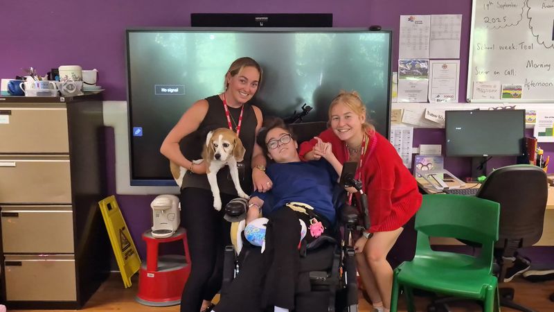 A lady with blonde hair is holding a beagle next to her daughter Lucie who is in a wheelchair. Next to Lucie is her carer in a red top, also named Lucy. They are standing in a classroom in front of a TV screen