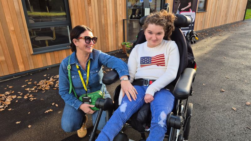 Mum Louise is crouched down next to her daughter Lilly who is in a wheelchair. A new building with timber cladding is behind them